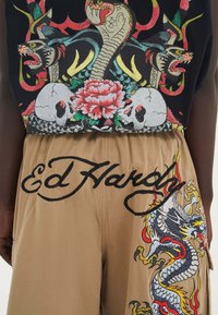 Černé grafické tričko s potiskem hadů, lebek a květin; béžové šortky s vyšitým designem draka a textem "Ed Hardy" v kurzivě.