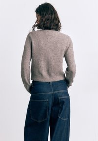 Kvinde med bølget brunt hår iført en grå striktrøje og mørkeblå viddebukser med høj talje, set bagfra mod en hvid baggrund.