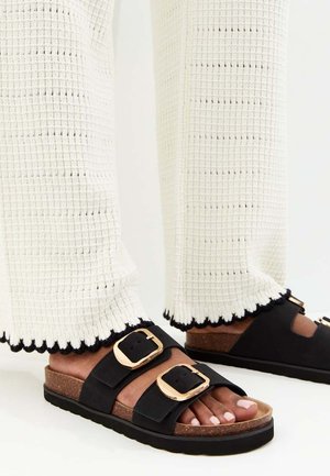 Piedi con sandali neri a ciabatta con grandi fibbie dorate e suole in sughero, abbinati a pantaloni bianchi con trama e orli bordati di nero.