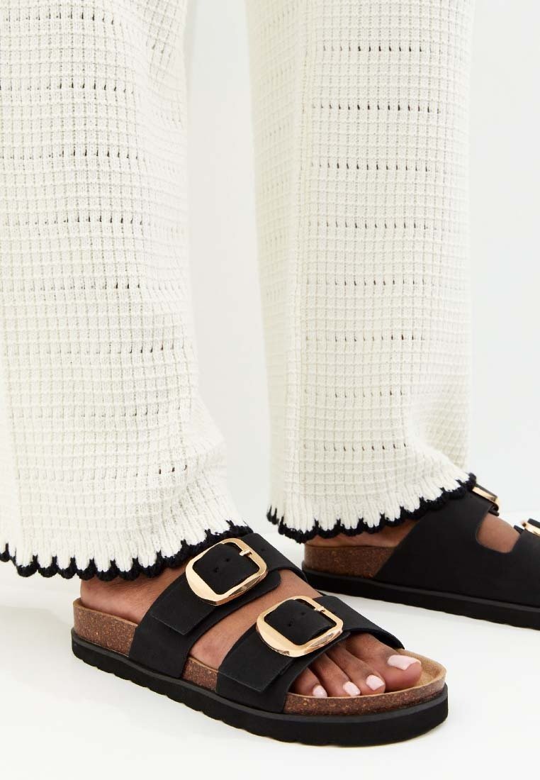 Pieds chaussés de sandales noires à brides avec de grandes boucles dorées et des semelles en liège, accompagnés d'un pantalon blanc texturé avec des ourlets bordés de noir.