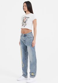 Hvid cropped t-shirt med grafisk print, parret med lyseblå baggy jeans med livlige gule accenter og en afslappet pasform. Hvide sneakers.
