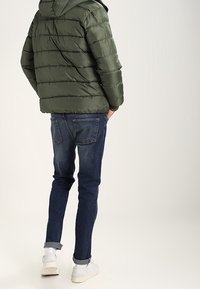 Grön puffjacka med horisontell quiltad mönster, parat med mörkblå skinny jeans och vita sneakers. Jackan har en huva.