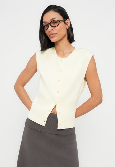 Cardigan sans manches en tricot jaune pâle avec un col rond et une fermeture à boutons sur le devant. Associé à une jupe côtelée gris foncé, taille haute.