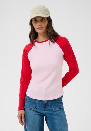 Jeune femme portant une casquette beige, un t-shirt rose à manches longues avec des manches rouges et un jean bleu, debout devant un fond uni.