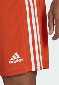 Orange Sportshorts aus leichtem Material haben drei weiße vertikale Streifen an der Seite und ein weiß gesticktes Logo am Saum.