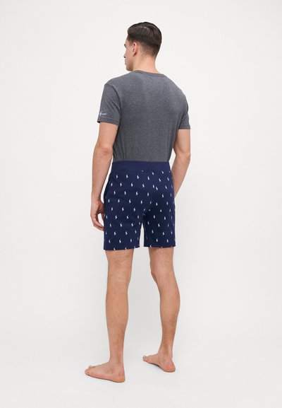 Shorts de bain navy avec motif de logo polo blanc, ceinture élastique, associés à une chemise grise à manches courtes. Personne de dos, pieds nus.