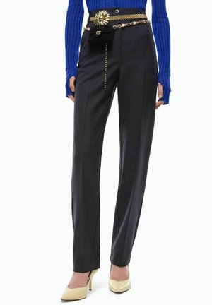 Mujer con pantalones negros a medida, suéter azul acanalado, cinturón de cadena dorada con detalle de flor y zapatos de tacón crema.
