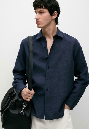 Mann, der ein dunkelblaues Hemd mit Knöpfen und weiße Hose trägt, eine schwarze Leder-Schultertasche hält und nach links schaut.