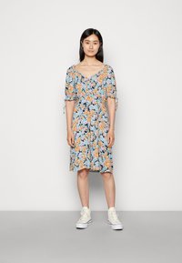 Vestido floral com um fundo escuro, apresentando flores laranja e azuis, mangas bufantes e detalhes com laço; combinado com ténis brancos.