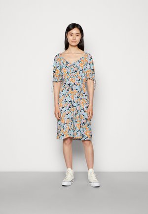 Vestido floral com um fundo escuro, apresentando flores laranja e azuis, mangas bufantes e detalhes com laço; combinado com ténis brancos.