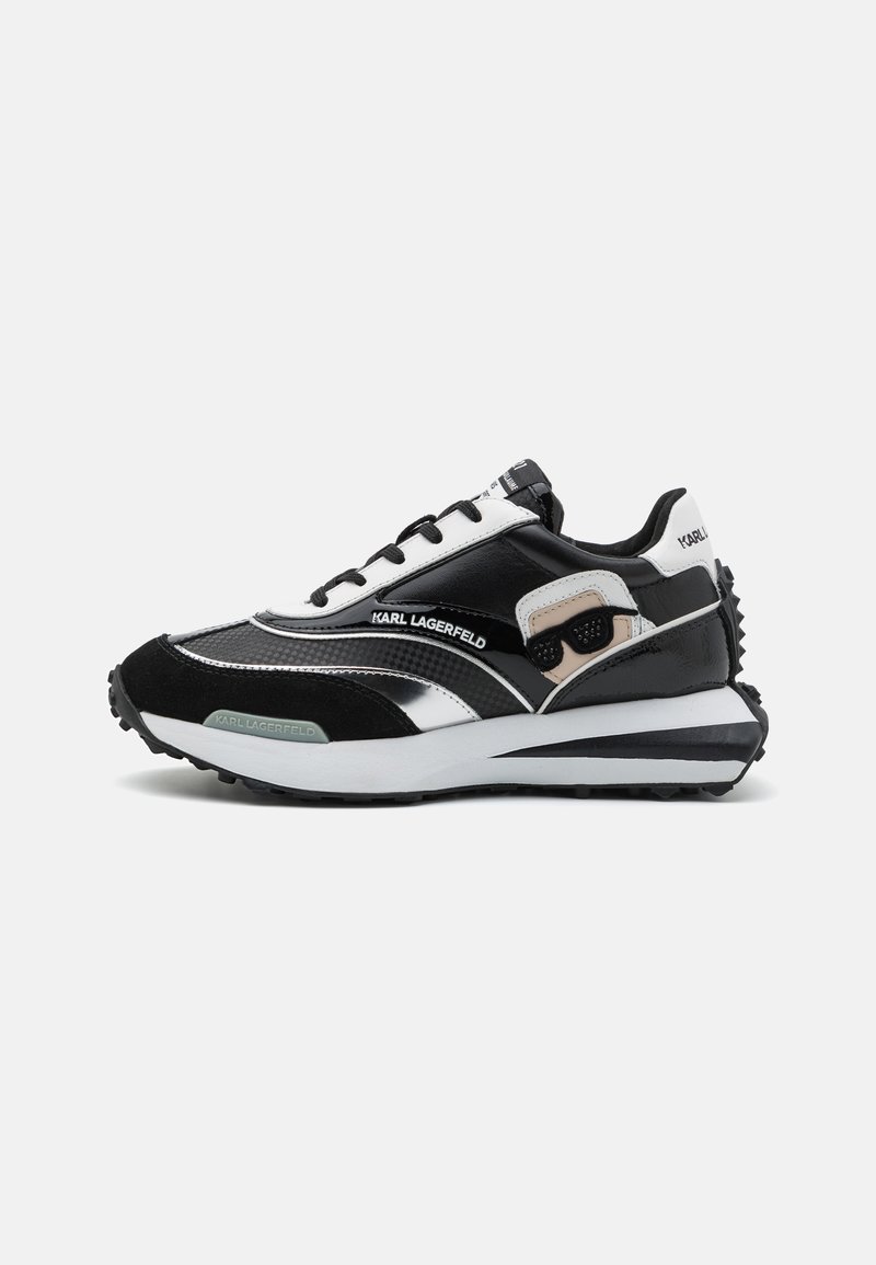 Fekete-fehér sneaker, amely velúrból és szintetikus anyagokból készült, reflektív díszítéssel, texturált panelek jellemzik, és vastag gumitalppal rendelkezik.