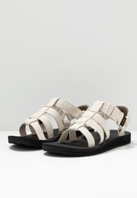 Beżowe sandały na paski z regulowaną klamrą, gumowymi podeszwami i czarnymi teksturowanymi wkładkami zapewniającymi przyczepność. Zaprojektowane z myślą o komforcie i codziennym użytkowaniu.