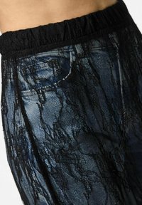 Pantaloni in pizzo nero con un motivo testurizzato, caratterizzati da una vita elastica e un tessuto stampato blu e nero sottostante.