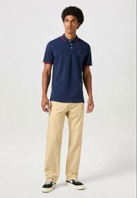 Camisa polo azul-marinho com detalhes em vermelho, mangas curtas e placket com dois botões, combinada com calças bejes claras e ténis pretos com solas brancas.