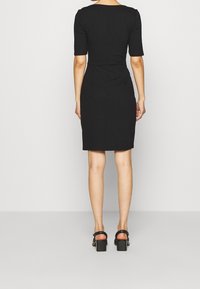 Robe noire ajustée à manches courtes, avec une longueur arrivant aux genoux et une texture lisse. Portée avec des sandales à talons noires sur un fond neutre.