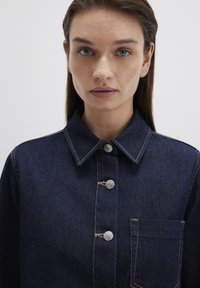 Dark blue denim jacket with a collar, silver buttons, and chest pocket. Features contrasting stitching and a textured fabric.