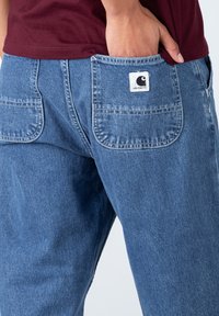 Pantaloni din denim albastru cu o croială relaxată, având două buzunare la spate și o etichetă de marcă pe buzunarul din dreapta. Material texturat cu detalii de cusătură.