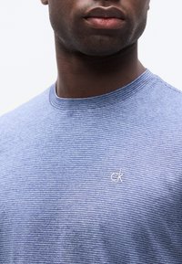 Κοντινό πλάνο ενός άνδρα που φοράει μπλε πουκάμισο με λεπτές ρίγες και μικρό λογότυπο "ck" στο στήθος, εμφανίζοντας το κάτω μέρος του προσώπου και τον λαιμό.