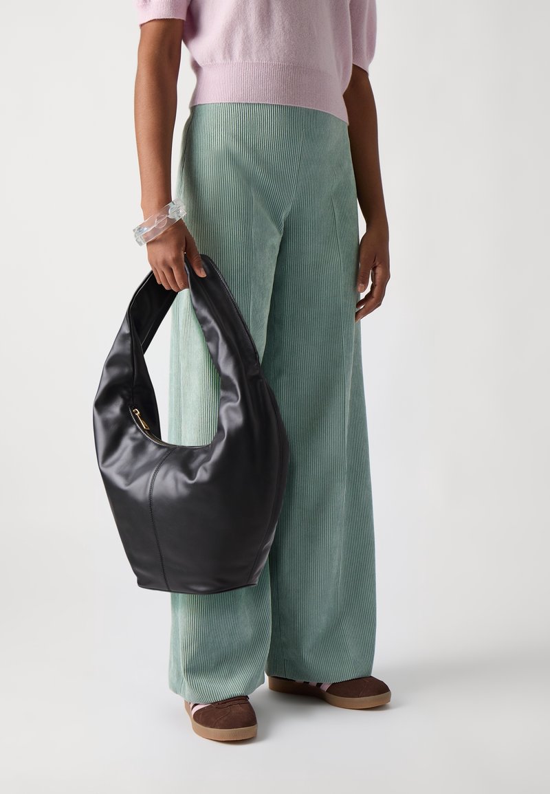 Personne portant un pantalon vert à jambes larges et un haut rose, tenant un grand sac hobo en cuir noir avec un bracelet transparent au poignet.