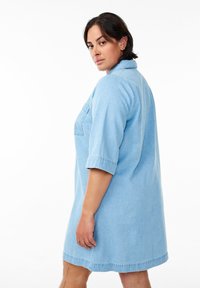 Šviesiai mėlynas džinsinis suknelė, trumpomis rankovėmis, su apykakle, viena krūtinės kišene ir laisvu siluetu, turinti tiesią apatinę dalį.