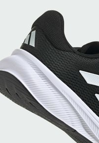 Schwarzer Sportschuh mit einem strukturierten Mesh-Obermaterial, weißer Zwischensohle und schwarzem Gummiaußensohle. Verfügt über reflektierende Akzente und ein aerodynamisches Design.