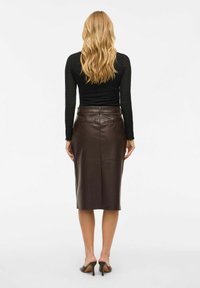 Vrouw met lang blond haar, gekleed in een zwart top met lange mouwen, een donkerbruin leren kokerrok en bruine hakken, met de rug naar de kijker gekeerd.