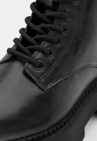 Stivale alla caviglia in pelle nera con una finitura liscia, punta rotonda, sei paia di occhielli e lacci neri su una suola in gomma spessa.