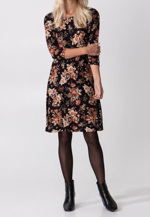 Femme portant une robe noire longueur genou imprimée de fleurs orange et beige, collants noirs transparents et bottines noires, debout les bras croisés.