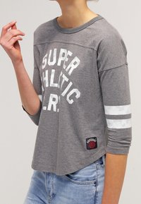 Grå sweatshirt med "SUPER ATHLETIC L.R." tryckt i vitt, trekvartsärmar, vita ränder och en svart och röd logotypapplikation vid fållen.