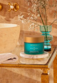 Turkossafärgad kruka med guldock lock märkt "The Ritual of Karma Whipped Body Cream" på en marmorbass. Mjuk handduk och en vas med blommor i närheten.