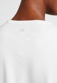 Detailní záběr na osobu v bílé tričku s kulatým výstřihem, na horní části zad pod výstřihem je malé šedé logo "CK".