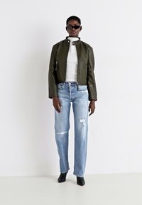 Chaqueta de cuero verde oliva, cuello alto blanco, vaqueros claros desgastados, botas negras con tacón y gafas de sol negras. Silueta moderna y sencilla.