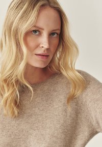 Svetle béžový svetr s jemným texturovaným pletem, vybavený kulatým výstřihem a mírně volným střihem. Modelka má vlnité blond vlasy.