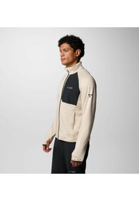 Giacca zip-up beige chiaro con tasca sul petto nera e logo. Presenta un tessuto texturizzato e un collo alto. Maniche lunghe con aperture per il pollice.