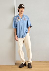 Ανοιχτό μπλε κοντομάνικο πουκάμισο με πολύχρωμες κεντητές λεπτομέρειες, λευκά κουμπιά, μπεζ παντελόνια και σκούρους καφέ μοκασίνες σε ξύλινο πάτωμα.