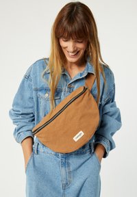 Sac en bandoulière en tissu marron avec une fermeture éclair, comportant une étiquette. Porté sur une veste en denim bleu clair, avec les mains du mannequin dans les poches.