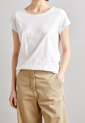Persoon draagt een simpel wit T-shirt met korte mouwen, ingestopt in beige hoge taillebroeken, met één hand in de zak.