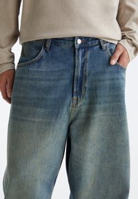 Homme portant un jean en denim bleu délavé et une chemise beige à manches longues, avec une main dans la poche et l'autre reposant sur le côté.