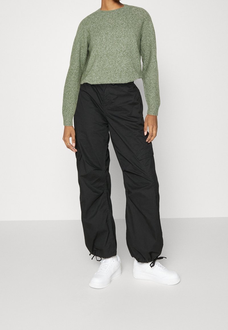 Personne portant un pull vert clair à manches longues, un pantalon cargo noir ample avec cordons aux ourlets et des baskets blanches, debout devant un fond uni.