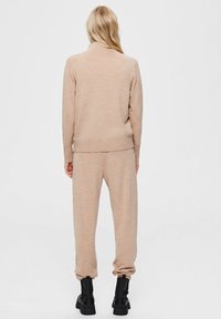 Béžový pletený set na domácí nošení, který obsahuje top s vysokým límcem a zúžené kalhoty. Materiál je měkký s vroubkovanými manžetami. Doplňeno černou obuví.