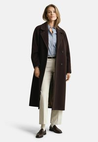 Langer dunkelbrauner Mantel mit großen Revers und doppelreihigen Knöpfen, getragen über einem hellblau gestreiften Hemd und cremefarbenen Hosen.