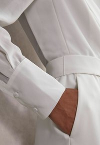 Balts garām piedurknēm krekls ar pogājamu aproci, kas izceļas ar gludu tekstūru un jostu viduklī, akcentējot elegantu dizainu un tīras līnijas.