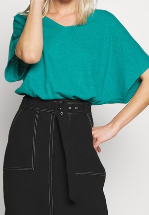 Kobieta ubrana w turkusową bluzkę z krótkim rękawem wpuszczoną w wysoką czarną spódnicę z białym przeszyciem i związanym paskiem.