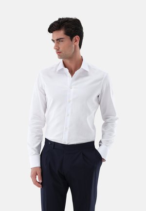 Camicia bianca con bottoni, maniche lunghe, colletto classico, tessuto liscio, abbinata a pantaloni blu scuro, design con piega singola.