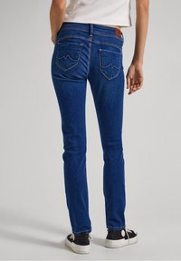 Blå denimjeans med slim fit, som har två bakfickor med ett vågmönster och en brun läderlapp vid midjan.