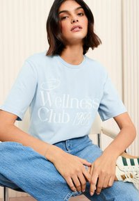 Gaiši zils kokvilnas t-krekls ar baltu drukājumu "The Wellness Club 1988". Īsas piedurknes un apaļa apkakle. Kombinēts ar ziliem džinsiem un sporta apaviem.