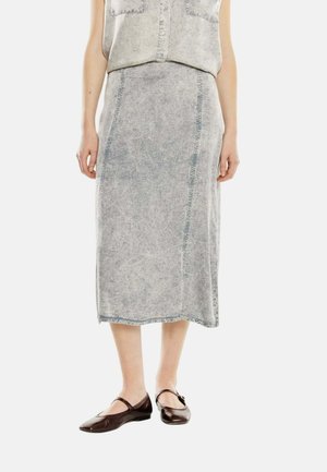 Mujer con falda midi gris descolorida combinada con un top sin mangas con botones y zapatos Mary Jane marrón oscuro, de pie sobre un fondo blanco.