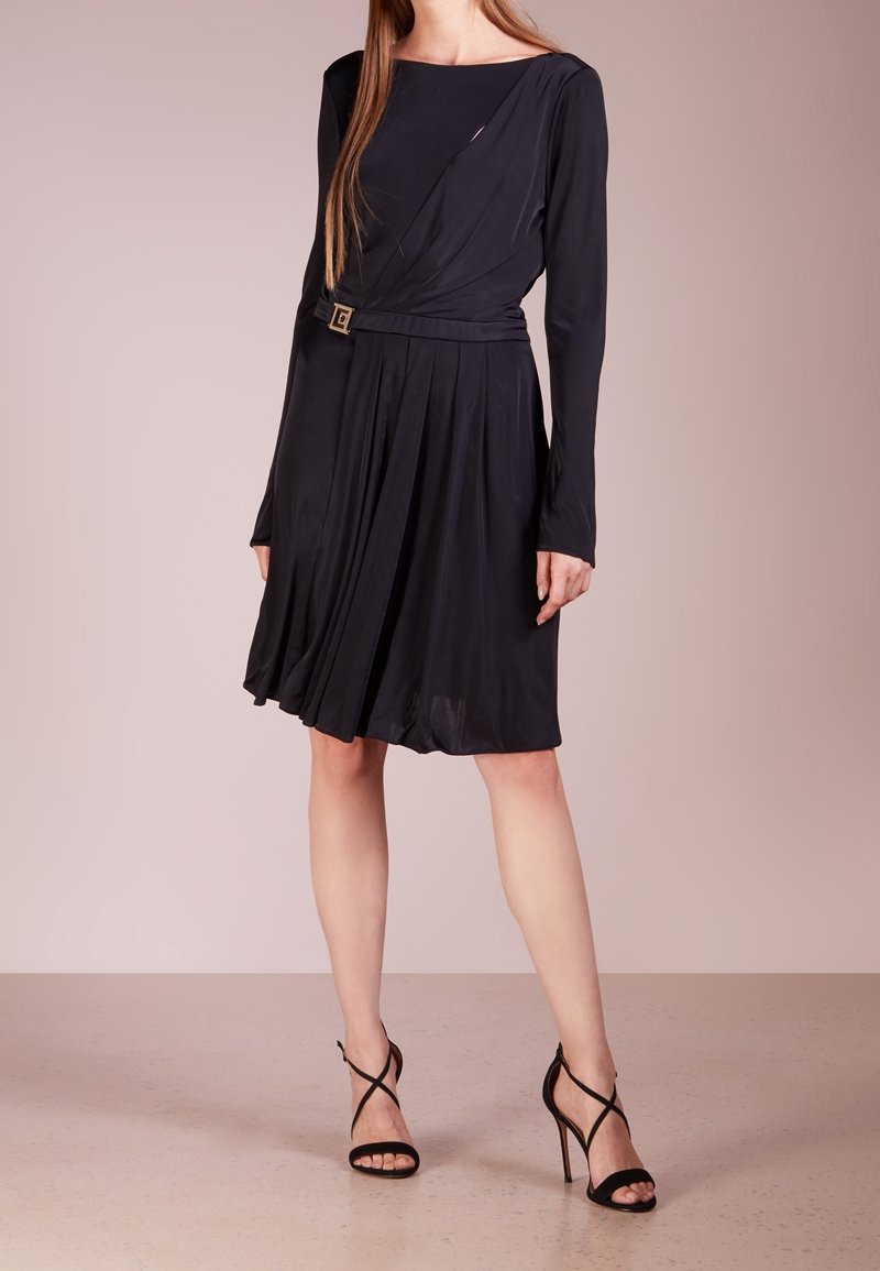 Robe noire à manches longues, décolleté carré et jupe plissée. Présente une ceinture accent en ton or à la taille. Associée à des talons noirs à lanières.