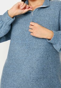 Pull bleu en polaire avec une fermeture éclair de quart de longueur, coupe décontractée et surface texturée. Présente des manches bouffantes et une apparence douce et chaude.