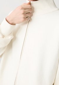 Weißes Zip-Jacke mit hohem Kragen, weichem, strukturiertem Stoff und schlankem Design. Mit sichtbarem Reißverschluss und lässig geschnittenen Ärmeln.